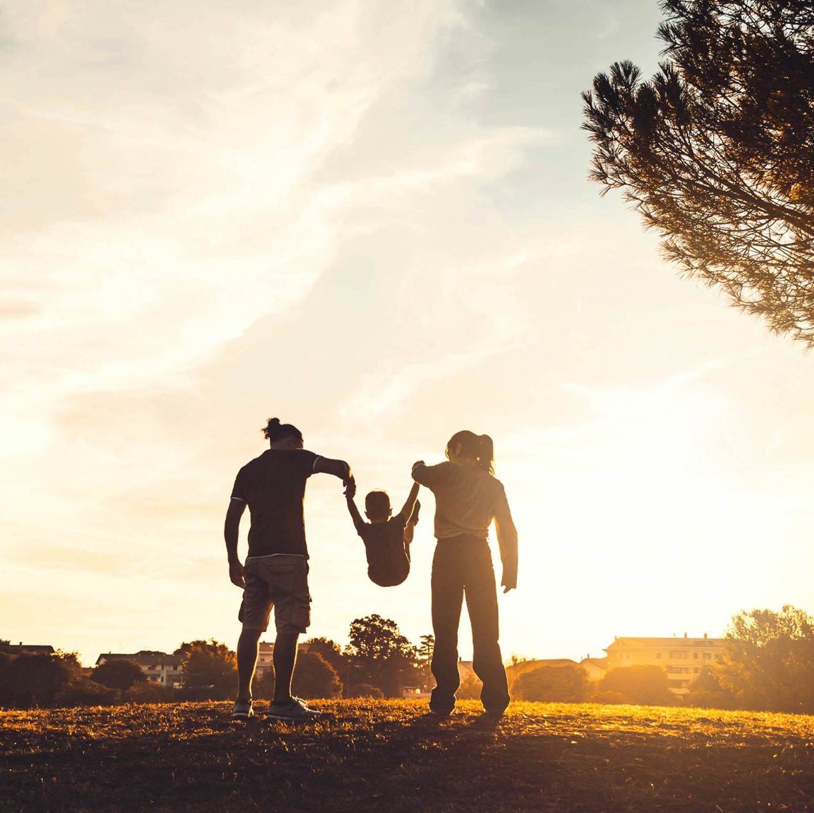 Deux personnes balancent leur enfant de manière ludique dans un beau parc avec un grand arbre et de l’herbe courte, au coucher du soleil.