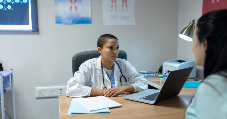 A doctor looks out over their desk at a patient, while a patient explains their situation.