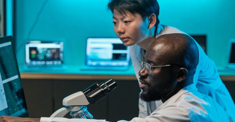 Two researchers stand over a desk with a microscope and computer screens, discussing something difficult.