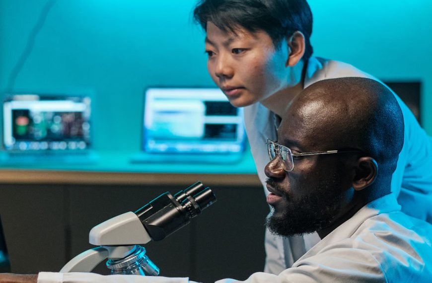 Two researchers stand over a desk with a microscope and computer screens, discussing something difficult.