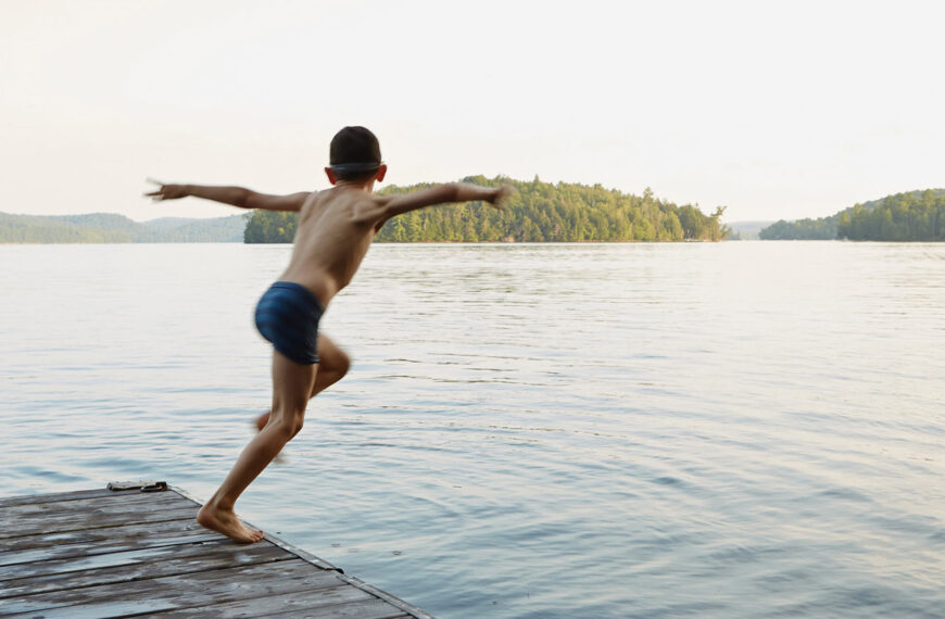 A young person gleefully runs and jumps off a dock into a stunning lake at a summer camp.