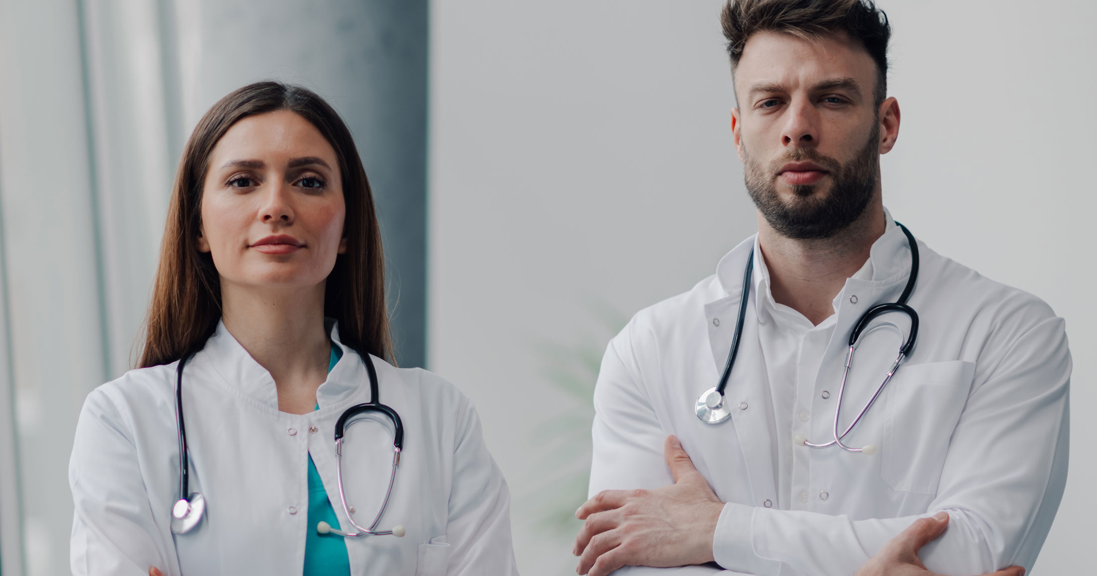 Two doctors stand shoulder to should looking at us in their white coats and stethoscopes.