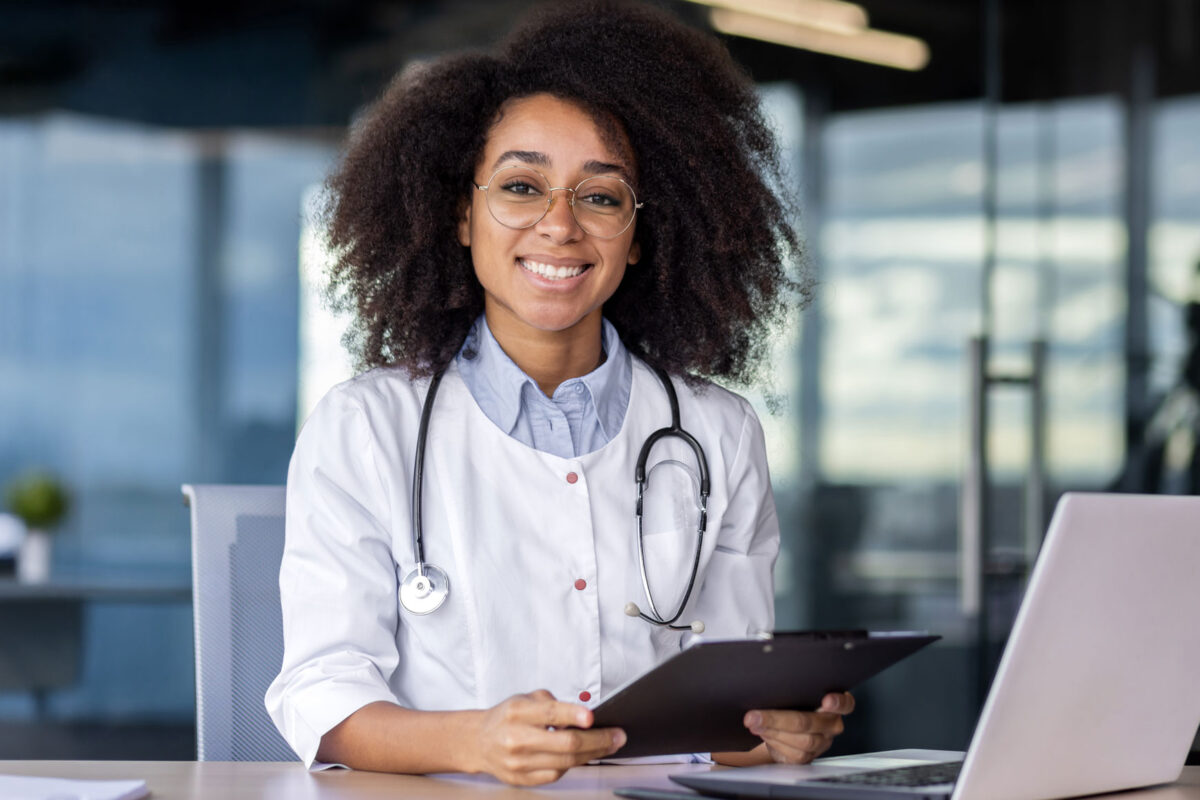 A doctor smiles sitting at their desk with a stethoscope around their neck.