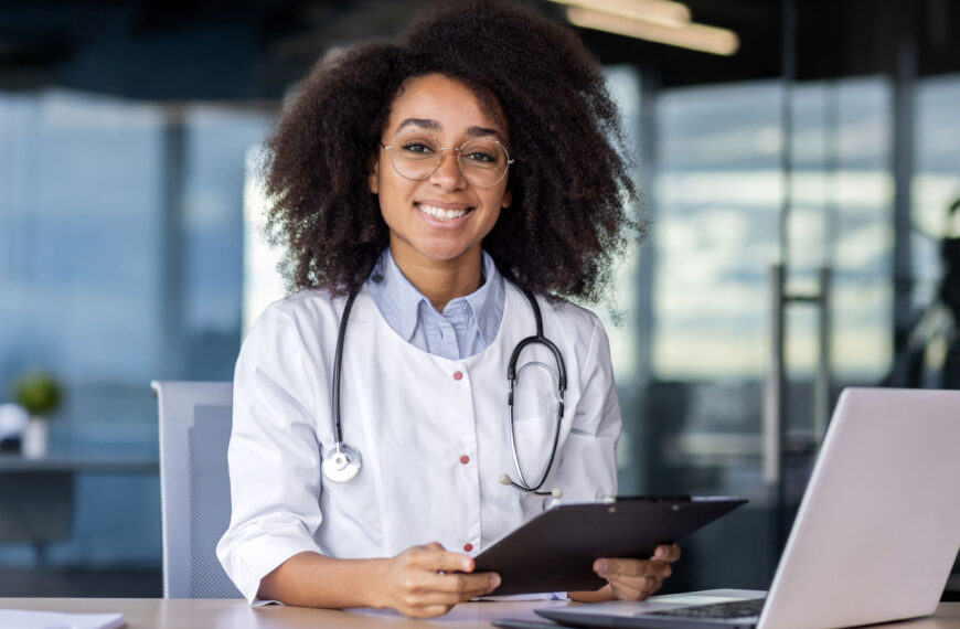 A doctor smiles sitting at their desk with a stethoscope around their neck.