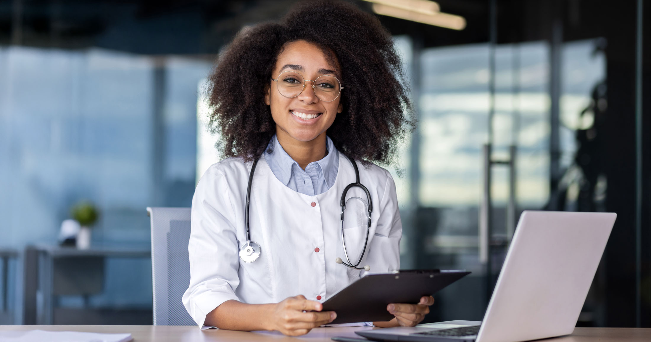 A doctor smiles sitting at their desk with a stethoscope around their neck.