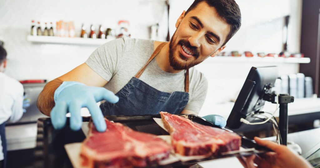 A butcher smiles while showing off some steaks over a counter.